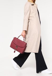 Bordowa torebka z klapką, złotymi akcesoriami i suwakiem, w zestawie z beżowym płaszczem i czarnymi spodniami, do tego białe trampki.