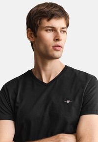 Mladý muž v čiernom tričku s véčkovým výstrihom a malým logom na hrudi, pozerá sa na stranu s neutrálnym výrazom, jednoduché svetlé pozadie.