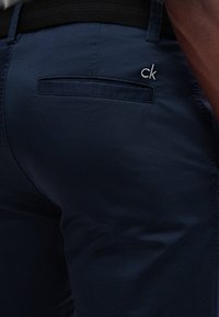 Gros plan sur un pantalon bleu marine avec une poche arrière et un petit logo blanc "ck" brodé au-dessus de la poche, porté avec une ceinture noire.