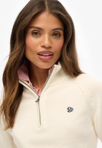 Cremefarbener Halbzip-Pullover aus strukturiertem Strickstoff, mit einem pinkfarbenen Innenkragen und einem kleinen dunkelblauen Logo auf der Brust.