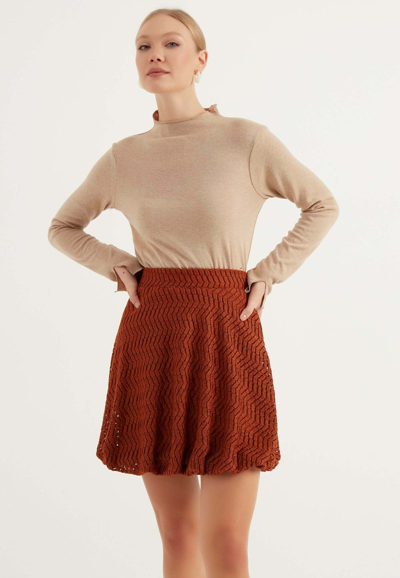Beige långärmad topp med hög hals, kombinerad med en rostfärgad kjol med zickzackmönster, som har en utställd form och texturerad väv.