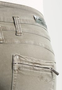 Grå stræk denim jeans med flere baglomme, herunder en lommer med lynlås. Har broderede detaljer og metalaccenter.