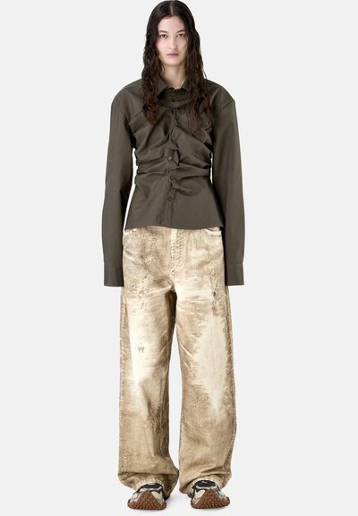 Persona joven con cabello largo y oscuro que lleva una camisa olivo arrugada abotonada, pantalones anchos beige desgastados y zapatos deportivos blancos con suelas marrones.