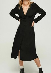 Abito midi nero con maniche lunghe, scollatura a V profonda e chiusura a bottoni davanti. Presenta dettagli arricciati in vita e uno spacco laterale. Tessuto liscio.