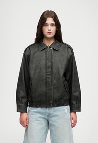 Czarna kurtka z imitacji skóry z kołnierzem, z przodu zapinana na napy, z bocznymi kieszeniami i elastycznym paskiem w talii. Nosi na sobie jasnoniebieskie dżinsy z przetarciami.