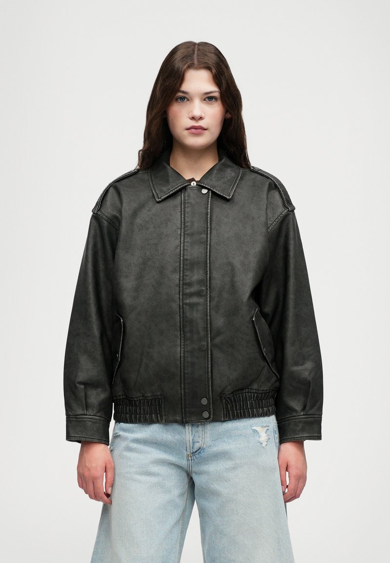 Czarna kurtka z imitacji skóry z kołnierzem, z przodu zapinana na napy, z bocznymi kieszeniami i elastycznym paskiem w talii. Nosi na sobie jasnoniebieskie dżinsy z przetarciami.