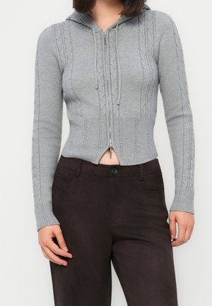 Person iført en tætsiddende grå kabelstrikket hættetrøje med lynlås, cropped kant og mørkebrune bukser mod en ensfarvet baggrund.