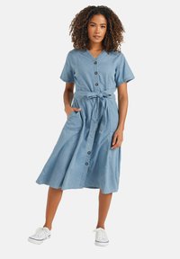 Vaaleansininen, polvipituinen chambray-mekko, jossa on lyhyet hihat, napitus edessä, sidottava vyö ja sivutaskut. Valkoiset tennarit viimeistelevät asun.