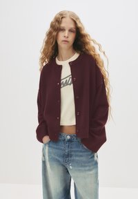 Cardigan borgogna con collo rotondo e chiusura a bottoni, indossato sopra una t-shirt cropped color crema. Presenta maniche ampie e polsini a coste. Jeans blu strappati.