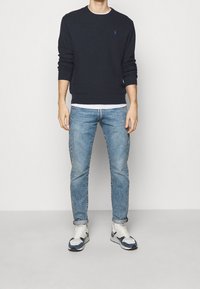 Mies, der einen dunkelblauen Pullover, hellblaue hochgekrempelte Jeans und weiße Turnschuhe trägt, steht vor einem einfarbigen Hintergrund.