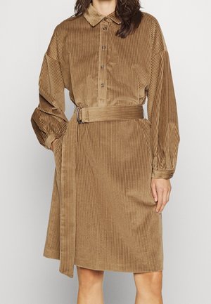 Kobieta ubrana w brązową sukienkę sztruksową do kolan z długimi rękawami, kołnierzykiem, zapinaną na guziki z przodu oraz z paskiem w talii w tym samym kolorze.