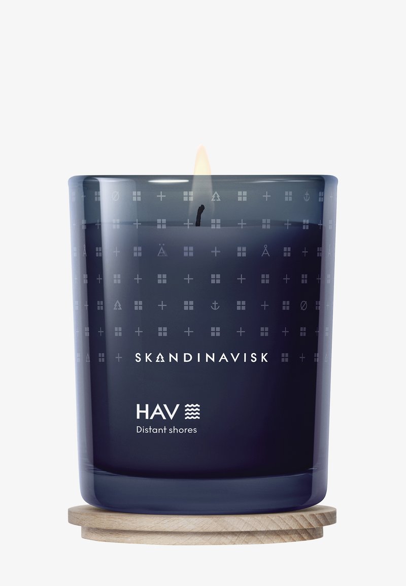 Ett mörkblått ljus i en glasbehållare med ingraverade mönster, med en träbas och en synlig låga. Etiketten lyder "SKANDINAVISK HAV Fjärran stränder."