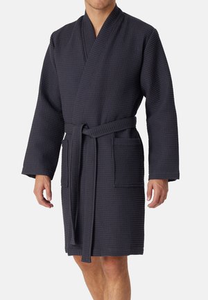 Mężczyzna ubrany w granatowy, sięgający do kolan szlafrok o fakturze wafla, z długimi rękawami, przednimi kieszeniami i paskiem związanym w talii.