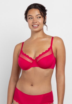 Vrouw draagt een felroze beugelbeha met mesh inzetstukken en bijpassende hoge taille slips, lacht en kijkt opzij.