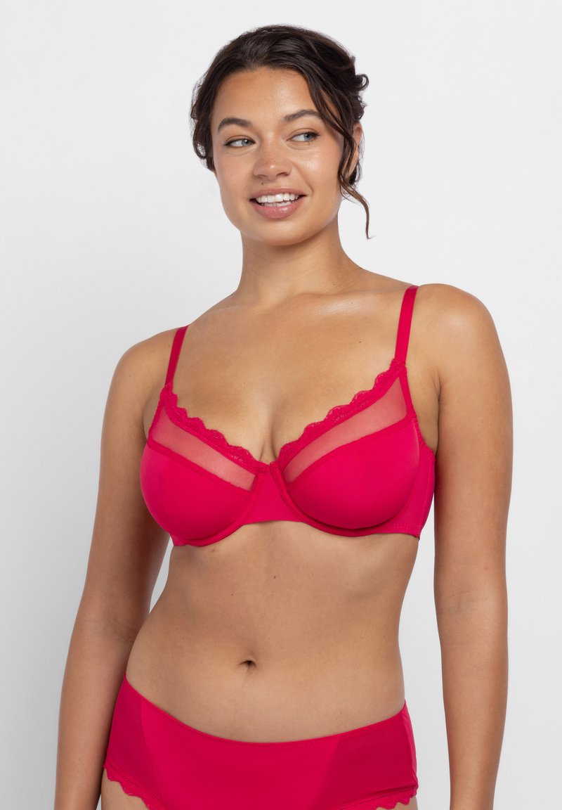 Žena oblečená v jasne ružovej podprsenke s kostícami a sieťovanými panelmi a zodpovedajúcich vysokých nohavičkách, usmievajúca sa a pozerajúca sa do strany.