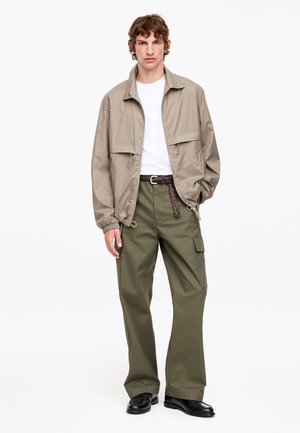 Jeune homme portant une veste légère beige, une chemise blanche, un pantalon large olive avec une ceinture tressée et des chaussures noires sur fond blanc.