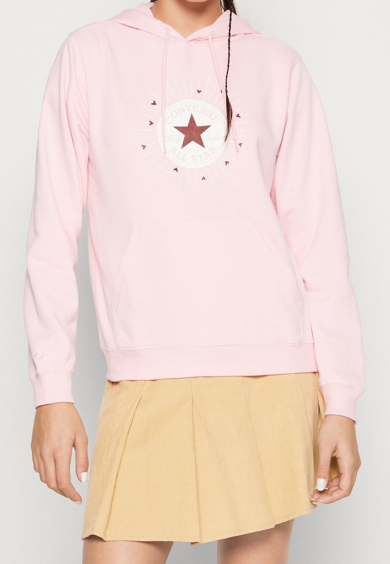 Femme portant un sweat à capuche Converse rose clair avec un logo étoile rouge et une jupe plissée beige, debout devant un fond simple.