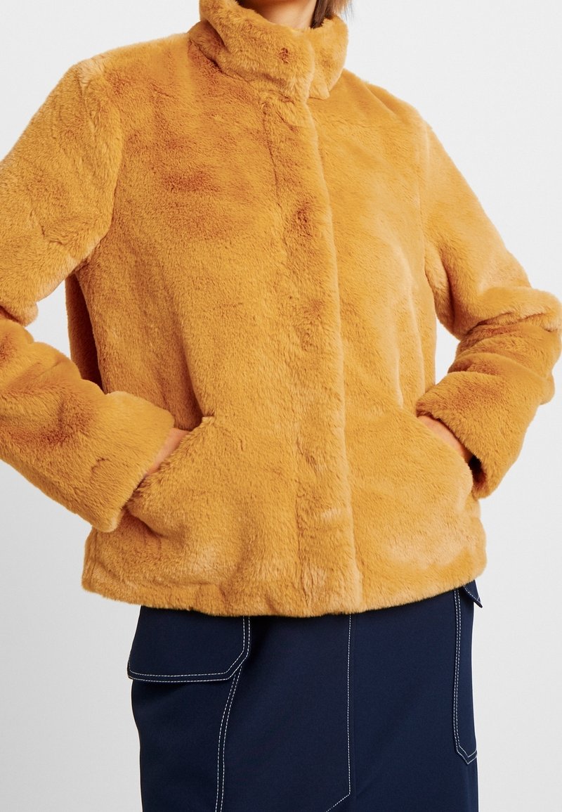 Kvinna iklädd en plysch, senapsgul fuskpälsjacka med händerna i sidofickorna och en marinblå kjol med vit sömnad.