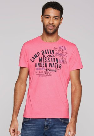 Mann trägt ein pinkes T-Shirt mit schwarzem und lila Text über Tauchen und Unterwassertouren, blaue Jeans, steht vor einem einfarbigen Hintergrund.