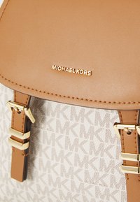 Tan leather backpack with gold hardware and a white patterned fabric featuring "MK" logos. Includes a magnetic flap and buckle accents.