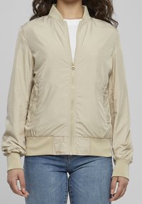 Beige bomberjacka med en slät nylonfinish, ribbade muddar och nederkant, dragkedjestängning och sidofickor. Klassisk, cropped skärning.