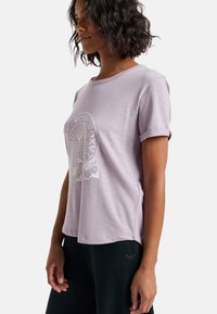 Camiseta corta de manga con un tono púrpura claro, con cuello redondo y puños enrollados. Presenta un gráfico blanco de flores y un logotipo en el frente.