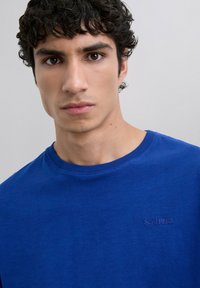Joven con cabello rizado y oscuro, vistiendo una camiseta azul básica de cuello redondo con un pequeño logo "SCALPERS" en el pecho, sobre un fondo liso.