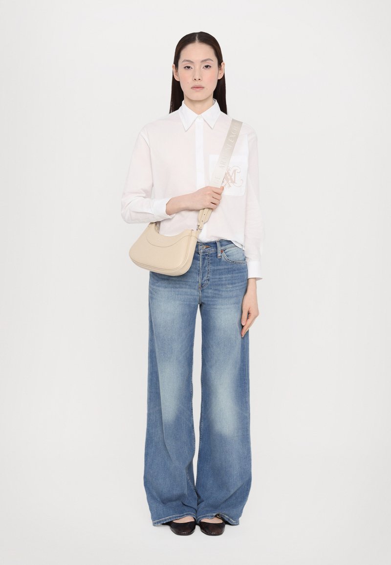 Borsa a spalla in pelle crema con design curvo, indossata a tracolla. Il modello indossa una camicia bianca con bottoni e jeans a gamba larga azzurri.