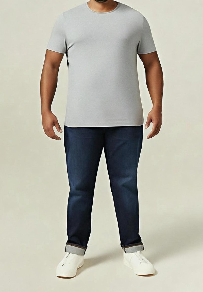Adulte portant un t-shirt gris clair à manches courtes, un jean bleu foncé retroussé et des baskets blanches, debout devant un fond uni.