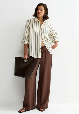 Vrouw met een wit gestreept shirt, bruine broek met brede pijpen, zwarte sandalen, die een grote donkerbruine handtas vasthoudt tegen een effen witte achtergrond.