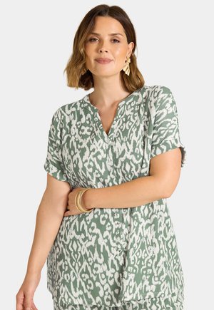 Femme aux cheveux bruns mi-longs portant de grandes boucles d'oreilles dorées et un haut boutonné à manches courtes à motifs verts et blancs avec des bracelets dorés.