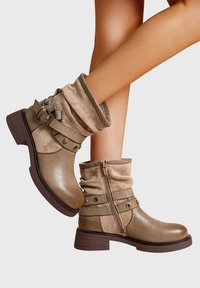 Bottes beiges à hauteur de cheville avec un dessus en daim froissé, des bandes décoratives et une fermeture éclair latérale, portées sur des jambes croisées devant un fond blanc.