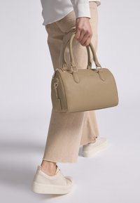 Beige leren handtas met een gestructureerde afwerking, ronde vorm, dubbele handvatten en hardware in goudtint; aangehechte tag met het logo van het merk.
