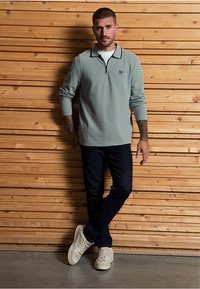 Světle zelený polo triko s dlouhým rukávem a zipem u krku, s tmavým lemováním, v kombinaci s tmavými džíny a bílými teniskami, s dřevěným pozadím.