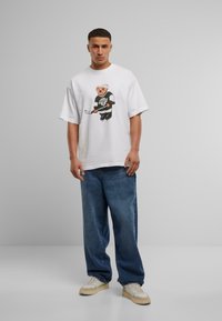 White cotton t-shirt with a graphic of a bear in a hockey jersey. Worn with baggy denim jeans and white sneakers, standing against a gray backdrop.
