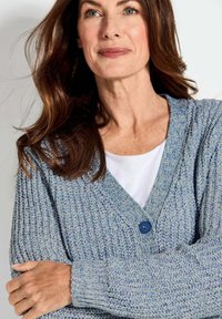 Lichtblauwe gebreide cardigan met een gestructureerd patroon, voorzien van een enkele blauwe knoop en een V-hals. Draagt over een witte top.