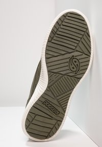 Semelle de basket kaki foncé avec des motifs de grip en caoutchouc texturé et un bord blanc. Logo "Dockers by Gerli" visible sur la semelle.