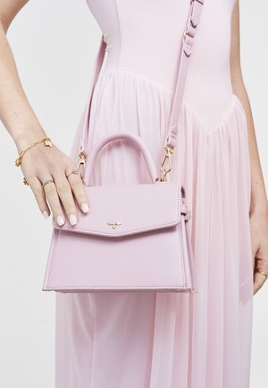 Žena v svetloružových šatách drží štruktúrovanú kabelku s vrchnou rukoväťou a zlatým detailom v tvare včely.