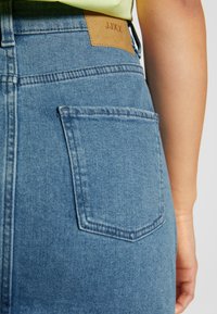 Saia de ganga azul com cintura alta, apresentando um único bolso traseiro e um remendo de couro bege com a inscrição "JJXX" na cintura.