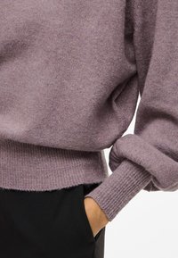 Vijolični pleteni pulover z ohlapnim krojem, rebrastimi manšetami in robom. Mehek material, vidne šive in črna oblačila z žepi pod njim.