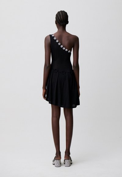 Donna con capelli intrecciati che indossa un vestito nero monospalla con dettagli floreali e scarpe con tacco a motivo bianco e nero, in piedi su sfondo grigio.
