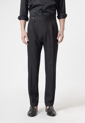 Hombre con pantalones negros a medida con pestaña en la cintura abotonada y bajos con puños, combinados con camisa negra y sandalias de cuero negras.