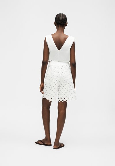 Γυναίκα με πλεξούδες, που φοράει λευκό αμάνικο τοπ με βαθύ V στο πίσω μέρος και λευκό σορτς με σχέδια, στέκεται σε λευκό φόντο.