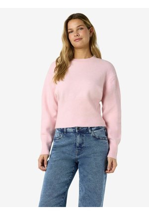 Femme aux longs cheveux ondulés portant un pull rose clair et un jean bleu taille haute, debout devant un fond blanc uni.