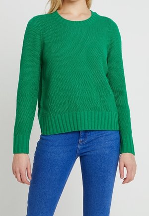 Femme portant un pull en maille texturée vert vif avec des poignets et un ourlet côtelés, associé à un jean en denim bleu, debout devant un fond blanc.