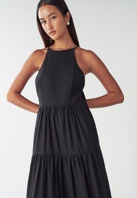 Žena s dlouhými tmavými vlasy má na sobě bezramínkovou černou šaty s volány a zlaté kruhové náušnice, pózuje s rukama za zády na bílém pozadí.