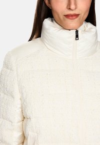 Weiße Daunenjacke mit strukturiertem gewebtem Stoff und glatten Nylonärmeln. Verfügt über einen hohen Kragen und einen Reißverschluss vorne.