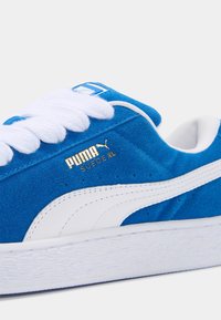 Baskets en daim bleu avec semelle en caoutchouc blanc. Elles sont dotées de lacets blancs et d'un logo doré sur le côté, mettant en avant une texture lisse et un design classique.