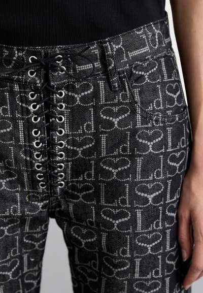 Pantalons en denim noir avec un devant lacé, œillets métalliques et un motif de logo répété en gris. Tissu texturé avec une coupe ajustée.