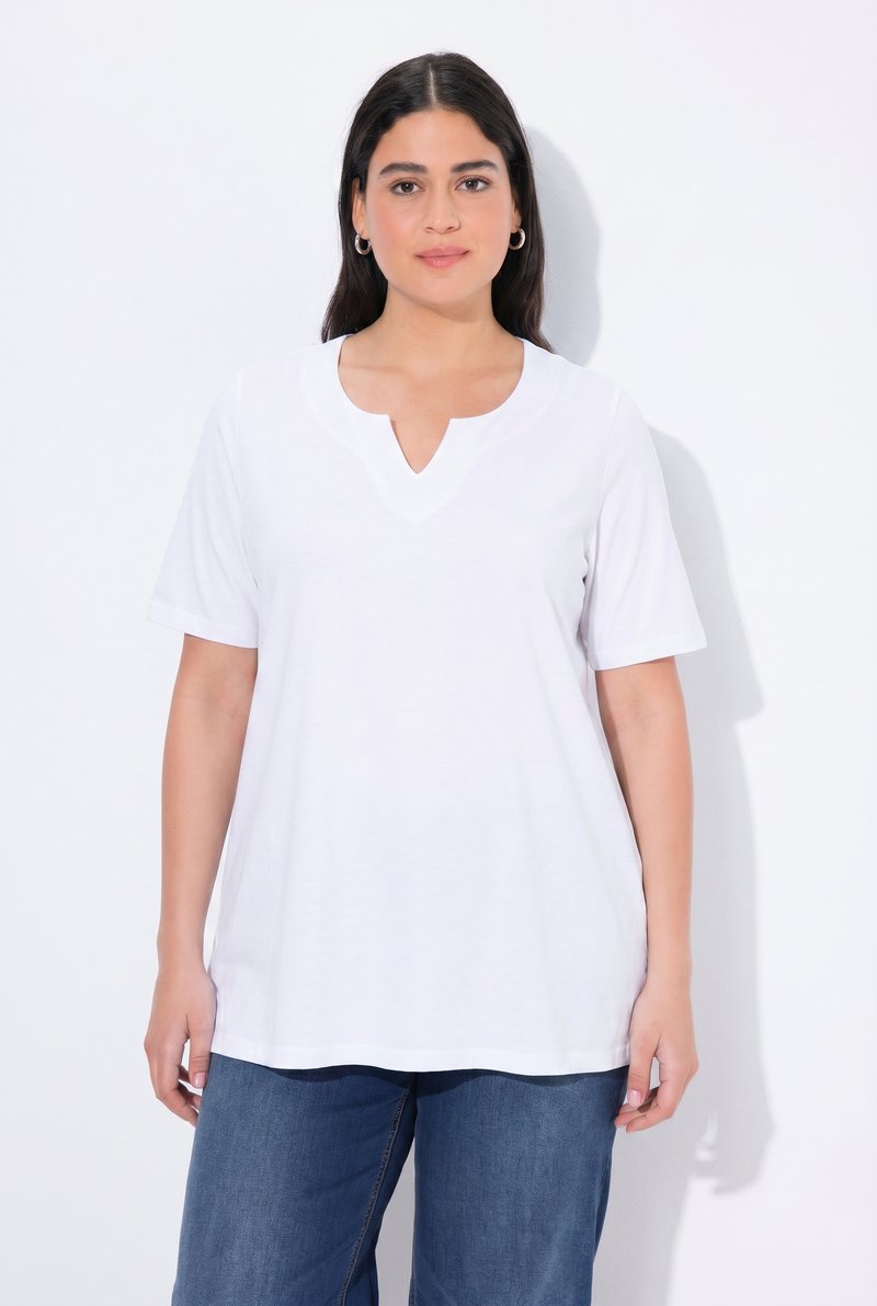 Femme aux longs cheveux foncés portant un t-shirt blanc uni à manches courtes avec un col en V et un jean bleu, debout devant un fond blanc.